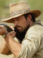 Close-up of Brandon Sklenar as Spencer Dutton in dark brown safari fedora aiming rifle in 1923 series outdoor scene