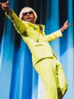 Tyler the Creator in lime green Igor suit performing live with arms spread wide blond wig and sunglasses on blue curtain stage