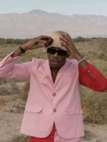 Tyler the Creator in pink Igor blazer with red trousers outdoors in desert landscape with blond wig and sunglasses
