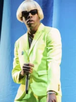 Tyler the Creator in lime Igor suit holding microphone with blond wig sunglasses and layered chain necklaces on blue curtain stage