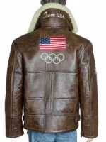 A close-up back view of the 2026 Winter Olympics Team USA brown leather flight jacket. The image focuses on the large American flag patch stitched onto the upper back panel, situated directly above the white embroidered Olympic rings logo, with a cream shearling collar peeking from above.
