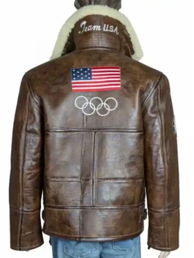 A close-up back view of the 2026 Winter Olympics Team USA brown leather flight jacket. The image focuses on the large American flag patch stitched onto the upper back panel, situated directly above the white embroidered Olympic rings logo, with a cream shearling collar peeking from above.