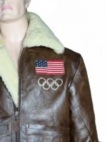 A close-up front view of the 2026 Winter Olympics Team USA brown leather flight jacket. The image focuses on the embroidered USA flag patch and white Olympic rings on the left chest, paired with a cream-colored plush shearling collar.