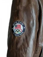 A close-up side view of the sleeve on the 2026 Winter Olympics Team USA brown leather flight jacket. The shot displays a circular navy blue embroidered patch with white text reading "UNITED STATES OLYMPIC TEAM", featuring an inner design of the American flag above the multi-colored Olympic rings.