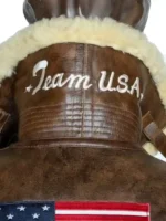 A close-up back view of the 2026 Winter Olympics Team USA brown leather flight jacket collar. The shot displays the white embroidered "Team USA" script text on the high-standing brown leather collar lined with cream shearling.