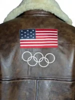 Back view of the 2026 Winter Olympics Team USA brown leather flight jacket on a mannequin. Features a large embroidered American flag patch across the upper back with the white embroidered Olympic rings below it. The high-standing leather collar shows white embroidered "Team USA" script text nestled into the reverse of the cream shearling trim, complemented by paneled accent stitching across the distressed brown leather back.