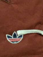 A close-up macro shot of the collar and upper chest of the Adidas x CLOT Fox Brown jacket, detailing the white embroidered Trefoil logo, traditional frog button knot, and textured twill fabric.