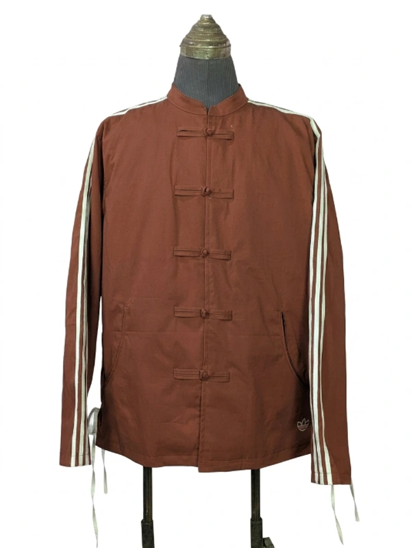 A studio product shot of a male model wearing the full-zip Adidas x CLOT Chinese New Year track jacket in Fox Brown, featuring a mandarin collar with traditional white frog closures over a silver zipper and white rope three-stripes down the sleeves.