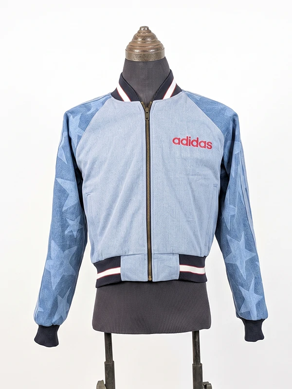 Complete front view of the full-zip Adidas Denim Graphic Varsity Bomber Jacket featuring a red embroidered logo, star-patterned sleeves, and a striped ribbed collar.