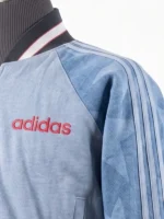 Close-up of the front chest of an Adidas denim varsity jacket featuring a red embroidered logo and raglan sleeves with signature three-stripe detailing.