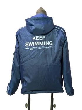 Full back view of the navy blue BTS Keep Swimming jacket. Features a large, bold white graphic reading "KEEP SWIMMING" across the upper back, underscored by a row of small, stylized light blue wave and swimmer illustrations just below the text.