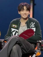 Jungkook seated smiling in black Kith varsity jacket showing KITH patch and Just Us embroidery at BTS Swimside 2026