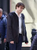 Robert Pattinson smiling in a black hooded jacket layered over a light shirt during an outdoor street scene