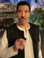 Lionel Richie wearing a black cotton vest and white shirt speaking while standing in an outdoor themed set