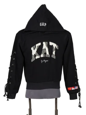 Front view of a black pullover fleece hoodie on a mannequin featuring large camo varsity lettering across the chest and decorative lace-up sleeve details.