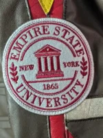 A detailed close-up shot of the circular Empire State University embroidered sleeve patch on the Peter Parker Photographer jacket, featuring red text and a university crest on a white background.