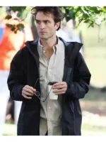 Robert Pattinson wearing a black hooded jacket over a light button shirt while holding glasses and a coffee cup outdoors