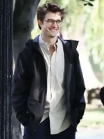 Robert Pattinson wearing glasses and a black hooded jacket over a light shirt in a bright outdoor setting