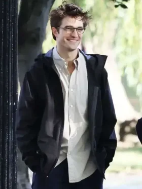 Robert Pattinson wearing glasses and a black hooded jacket over a light shirt in a bright outdoor setting