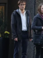 Robert Pattinson in a black hooded jacket with a white shirt and dark pants, standing outside near a building entrance