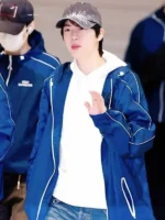 BTS member wearing blue Keep Swimming windbreaker jacket over white hoodie at airport