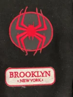 Close-up of Miles Morales varsity jacket front patches showing circular red spider symbol badge and Brooklyn New York rectangular patch on black wool body