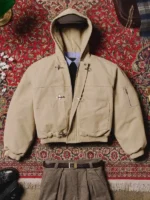 Beige Shinzo Shige canvas clasp bomber jacket styled with belt on ornate red Persian rug background