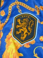 Close-up of the woven black and gold KNVB shield patch on the blue fabric of the FIFA World Cup 2026 Netherlands Anthem Jacket.