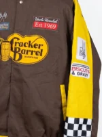 Extreme close-up of the left sleeve and torso of the Cracker Barrel 400 racing jacket, showcasing the yellow sleeve panel with embroidered patches for a rocking chair and "Biscuits & Gravy" over a checkered cuff pattern.
