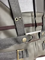 Macro close-up shot of the front brass buckle closure, dark green leather texture, and canvas harness straps with brass studs on the Bane vest.
